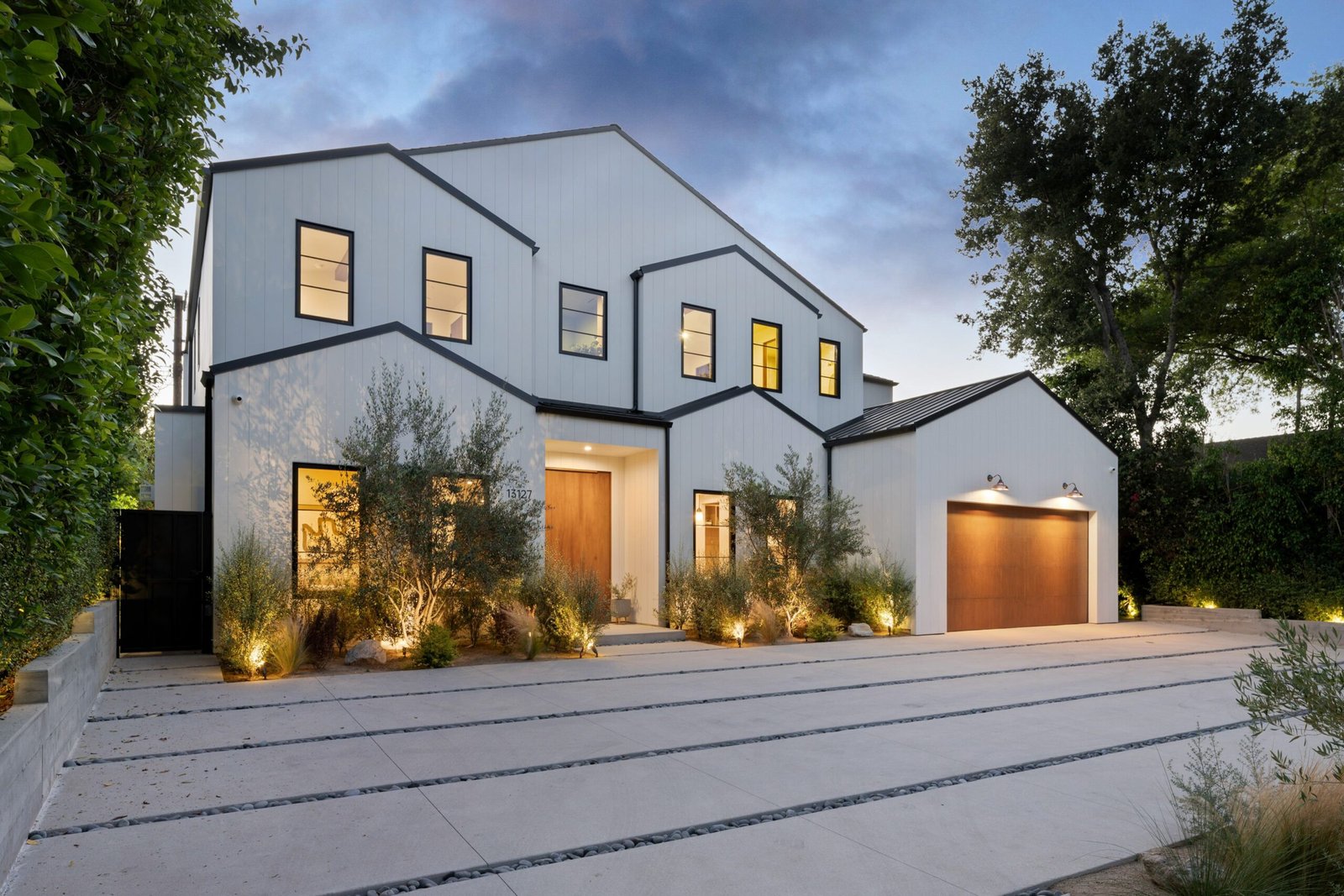 Modern two-story house with illuminated windows and landscaped driveway at dusk, surrounded by greenery.